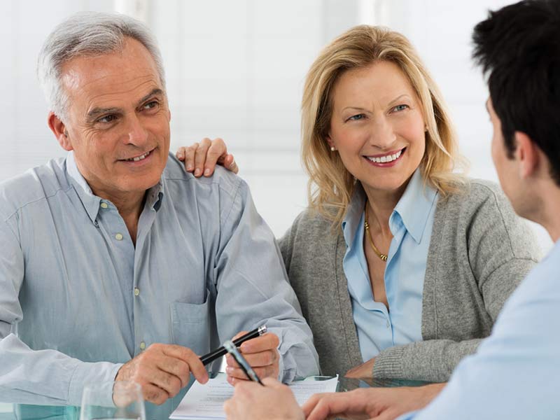An older couple is smiling and engaging in a conversation with a younger individual who is writing on a document, likely in a professional consultation or meeting setup.